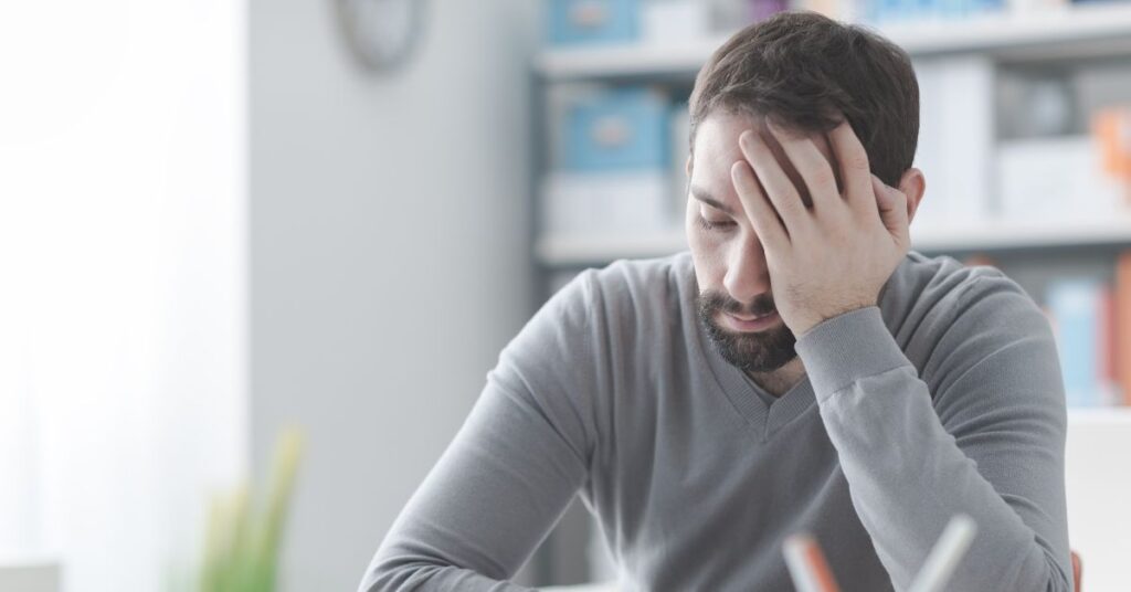 Stressed and exhausted man with his head in his hand, sitting at a desk in an office, symbolizing burnout and fatigue from work