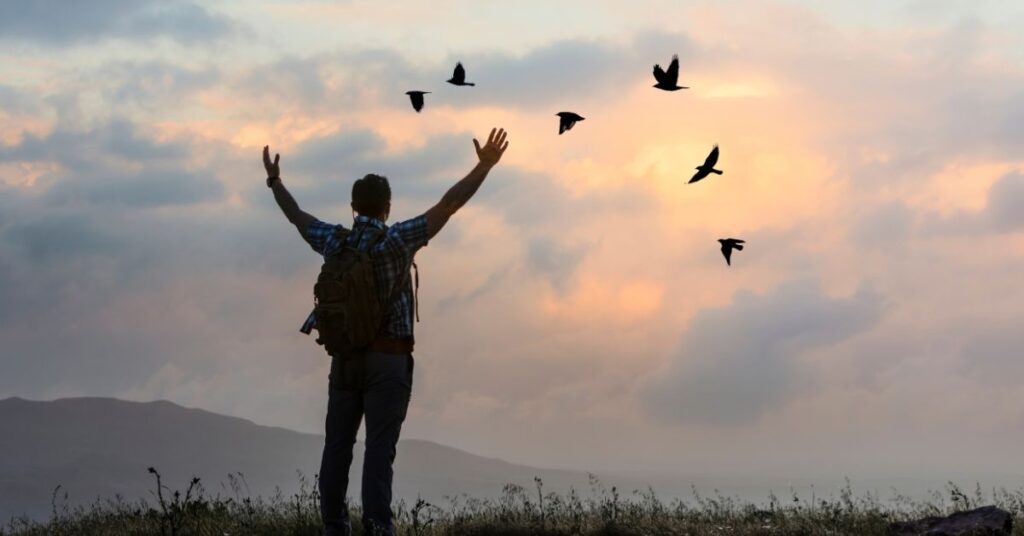 Person with arms raised standing on a hilltop at sunrise, symbolizing freedom and breaking free from constraints, with birds flying in the sky.